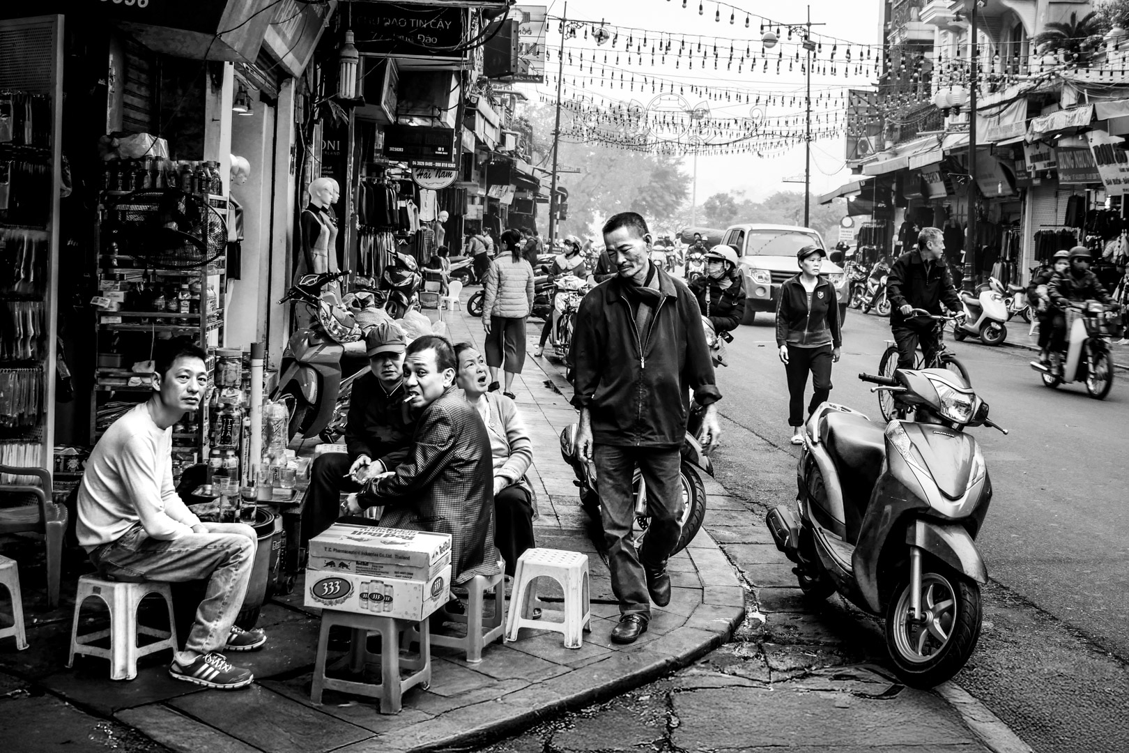 Black and white street photography of people sitting on plastic stools and motorcycles passing by on a busy street in Vietnam, capturing urban social culture